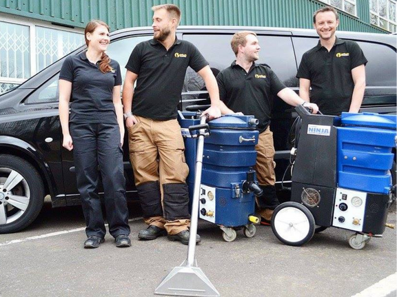 This is a photo of Ruislip Carpet Cleaning Solutions carpet cleaners (three men and one woman) standing in fromt of their black van, with two steam cleaning carpet machines next to them